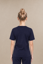 A woman viewed from the back wearing a navy blue underscrub set consisting of a short-sleeve underscrub top and matching pants, standing against a plain beige studio background.