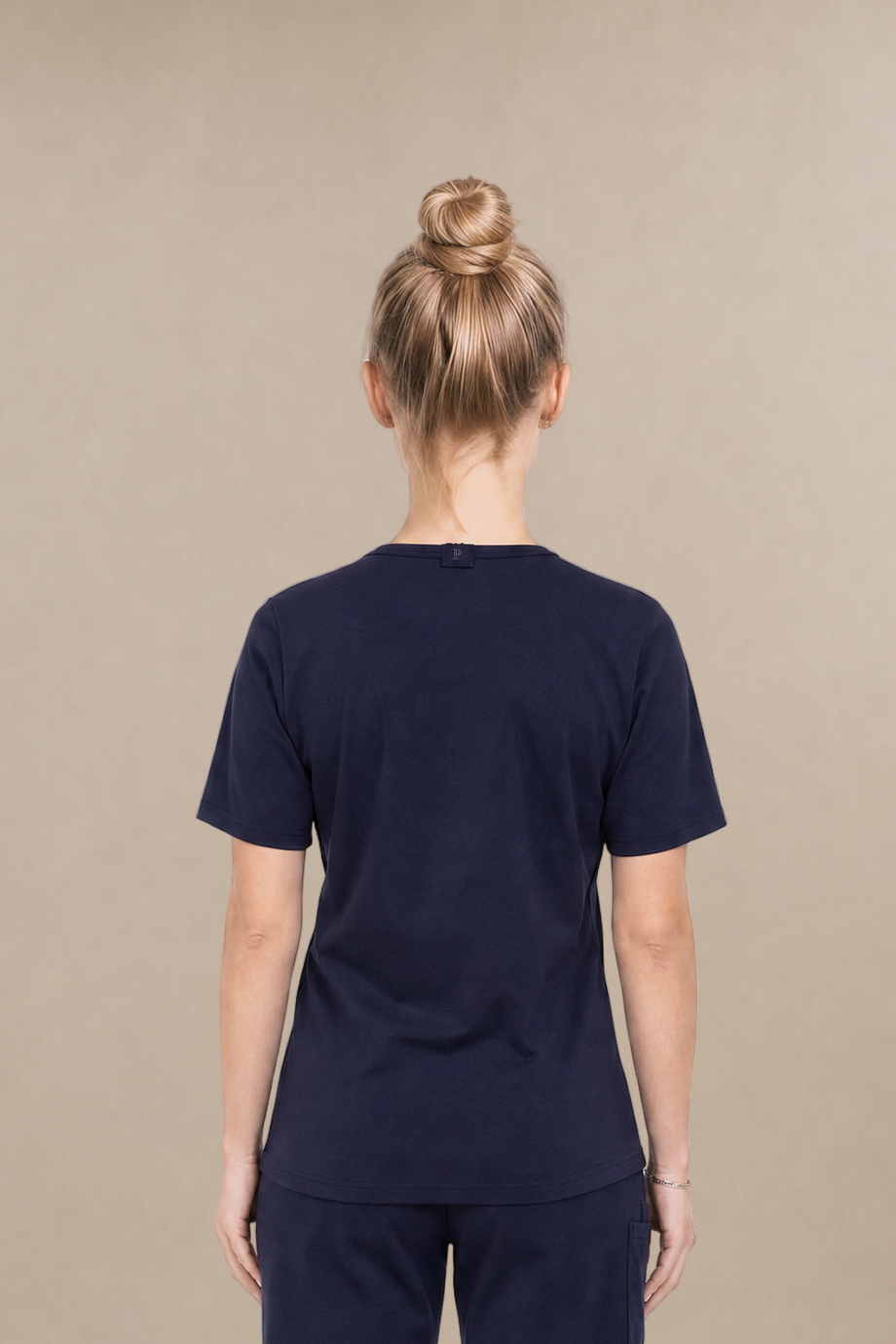 A woman viewed from the back wearing a navy blue scrub set consisting of a short-sleeve underscrub top and matching pants, standing against a plain beige studio background.