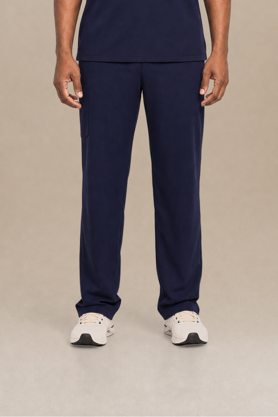 A man modeling a navy blue V-neck scrub top and matching scrub pants set, standing against a neutral beige studio background.