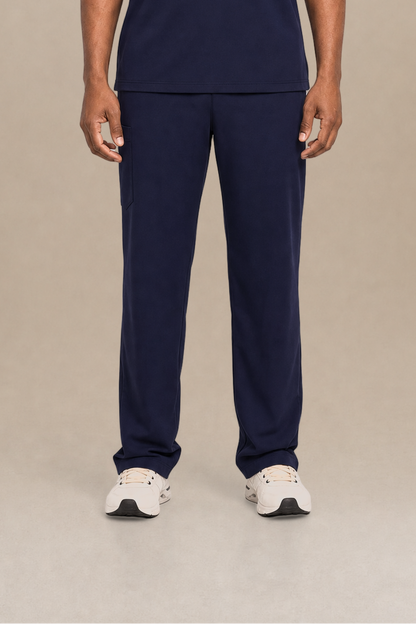 A man modeling a navy blue V-neck scrub top and matching scrub pants set, standing against a neutral beige studio background.