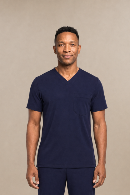 A man modeling a navy blue V-neck scrub top and matching scrub pants set, standing against a neutral beige studio background.