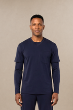 A man modeling a matching navy blue long-sleeve underscrub shirt layered under a navy blue short-sleeve scrub top and scrub pants set, standing against a solid beige studio background.