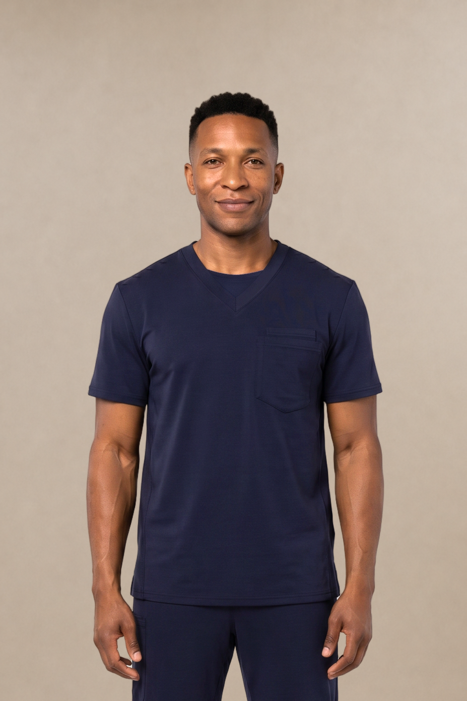 A man modeling a navy blue short-sleeve crewneck scrub shirt and matching navy blue scrub pants, standing against a solid beige studio background.
