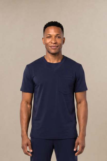 A man modeling a navy blue short-sleeve crewneck scrub shirt and matching navy blue scrub pants, standing against a solid beige studio background.