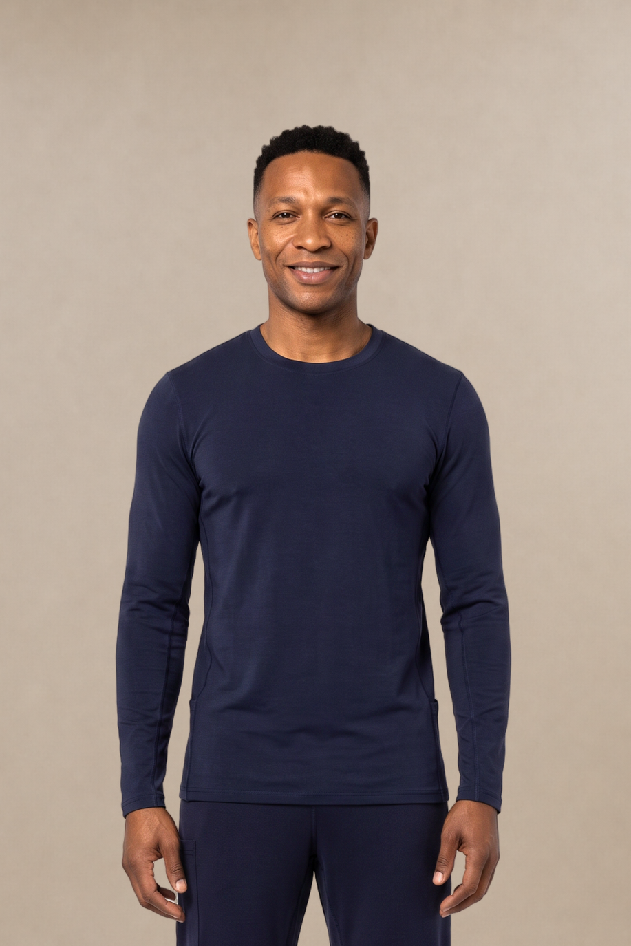 A man modeling a navy blue long-sleeve crewneck underscrub shirt and matching navy blue pants against a solid beige studio background.