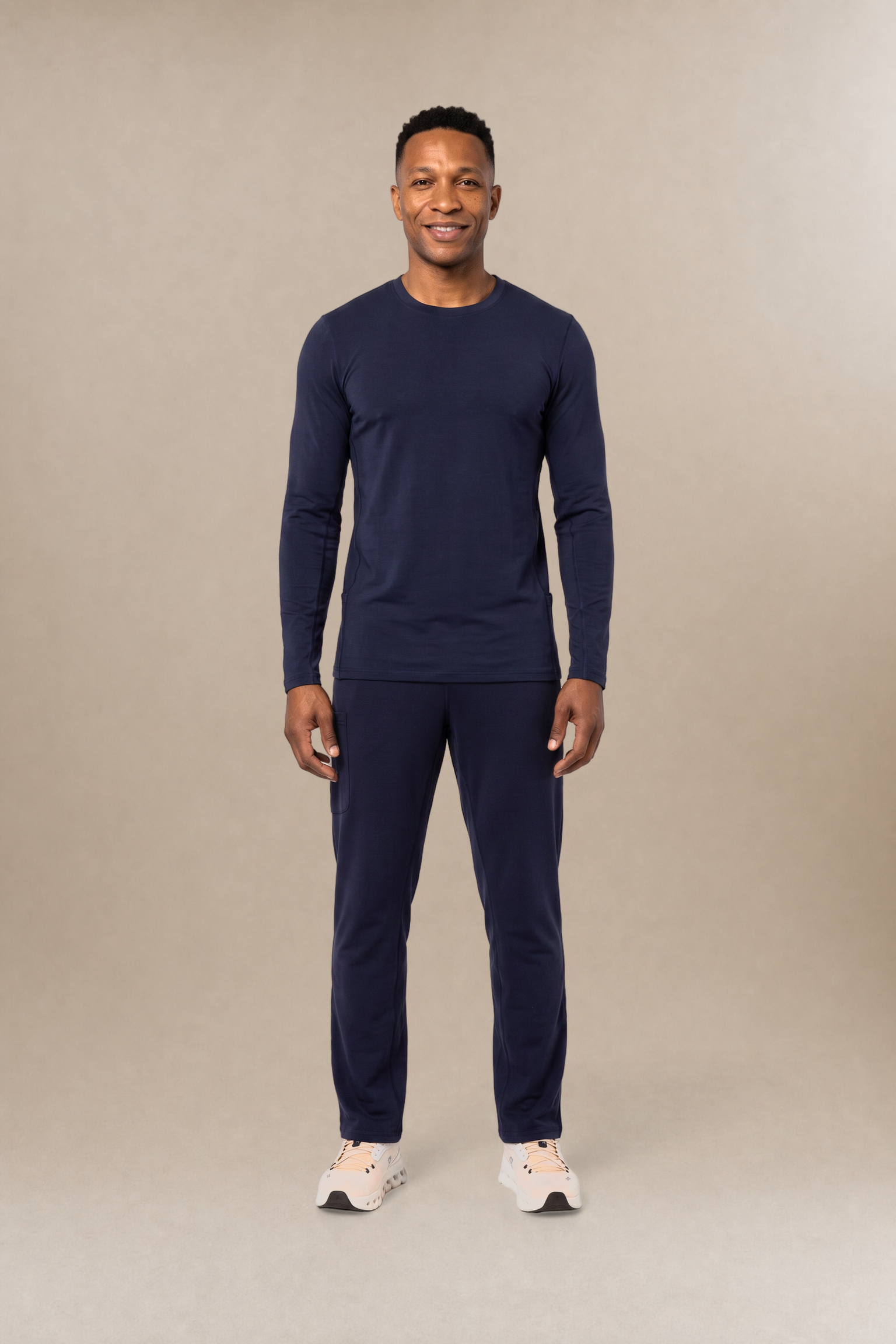 A man modeling a navy blue long-sleeve crewneck underscrub shirt and matching navy blue pants against a solid beige studio background.