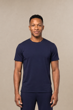 A man modeling a matching navy blue short-sleeve underscrub shirt and scrub pants set, standing against a solid beige studio background.
