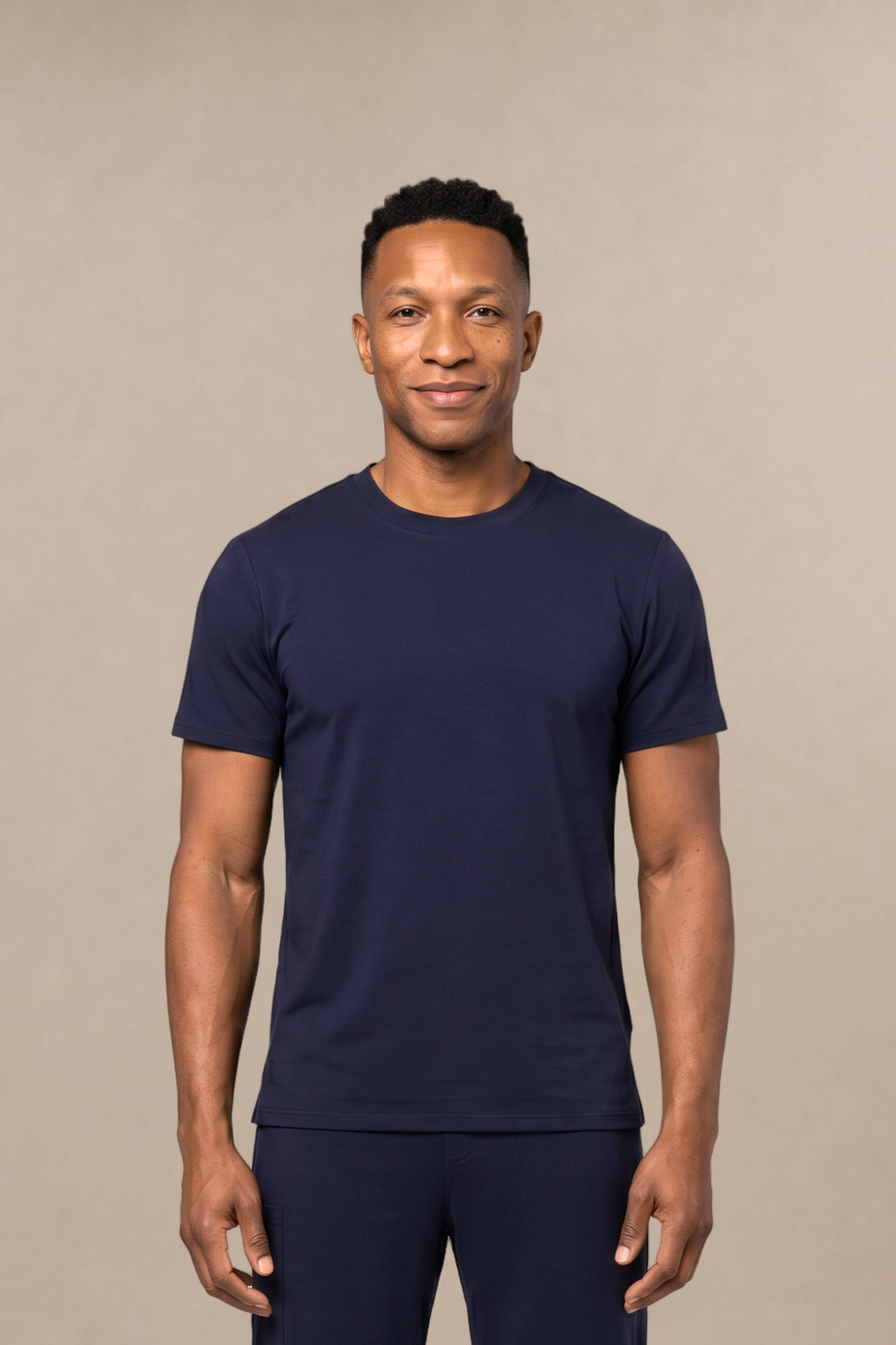 A man modeling a matching navy blue short-sleeve underscrub shirt and scrub pants set, standing against a solid beige studio background.