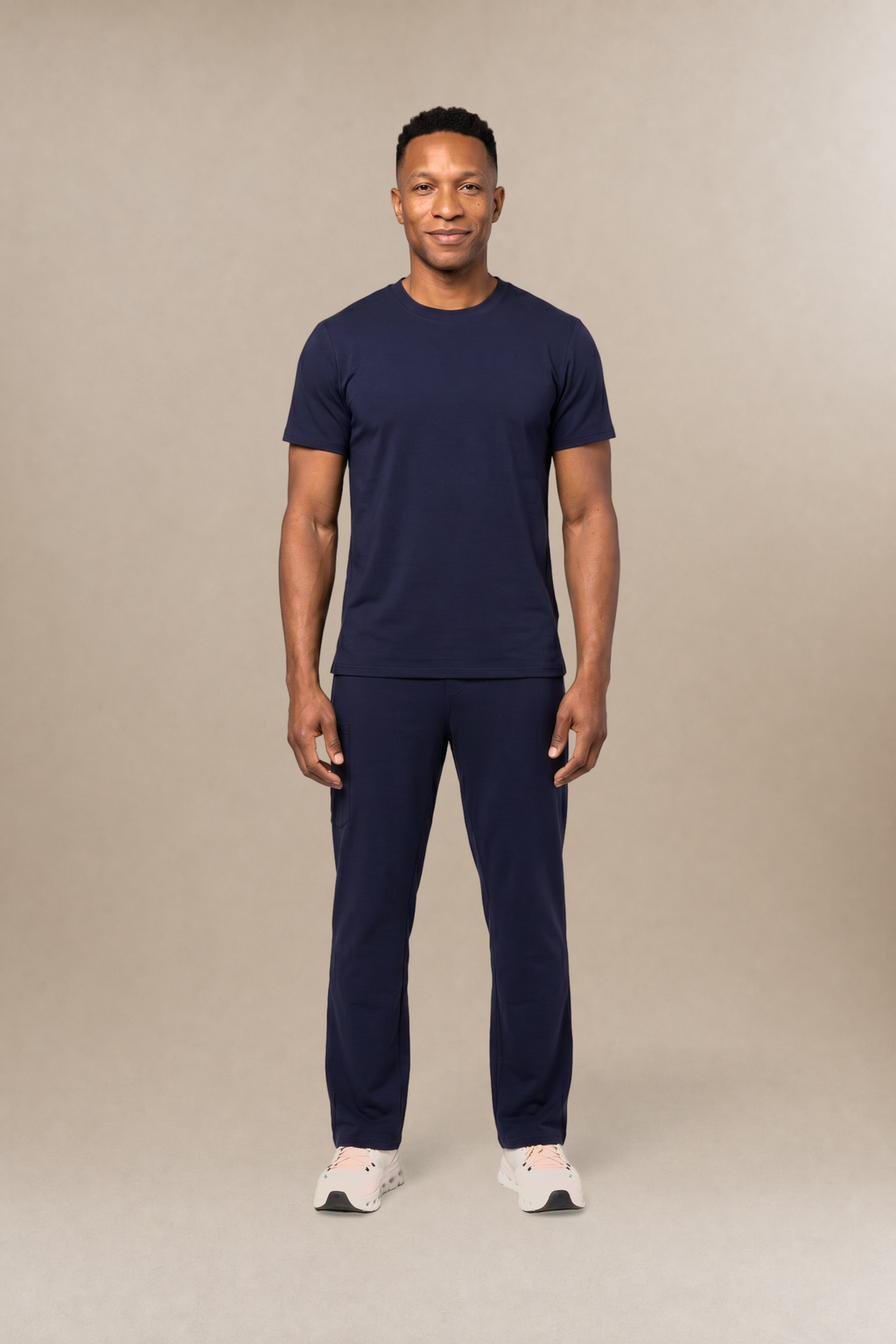 A man modeling a matching navy blue short-sleeve underscrub shirt and scrub pants set, standing against a solid beige studio background.