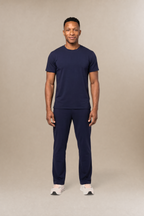 A man modeling a matching navy blue short-sleeve underscrub shirt and scrub pants set, standing against a solid beige studio background.