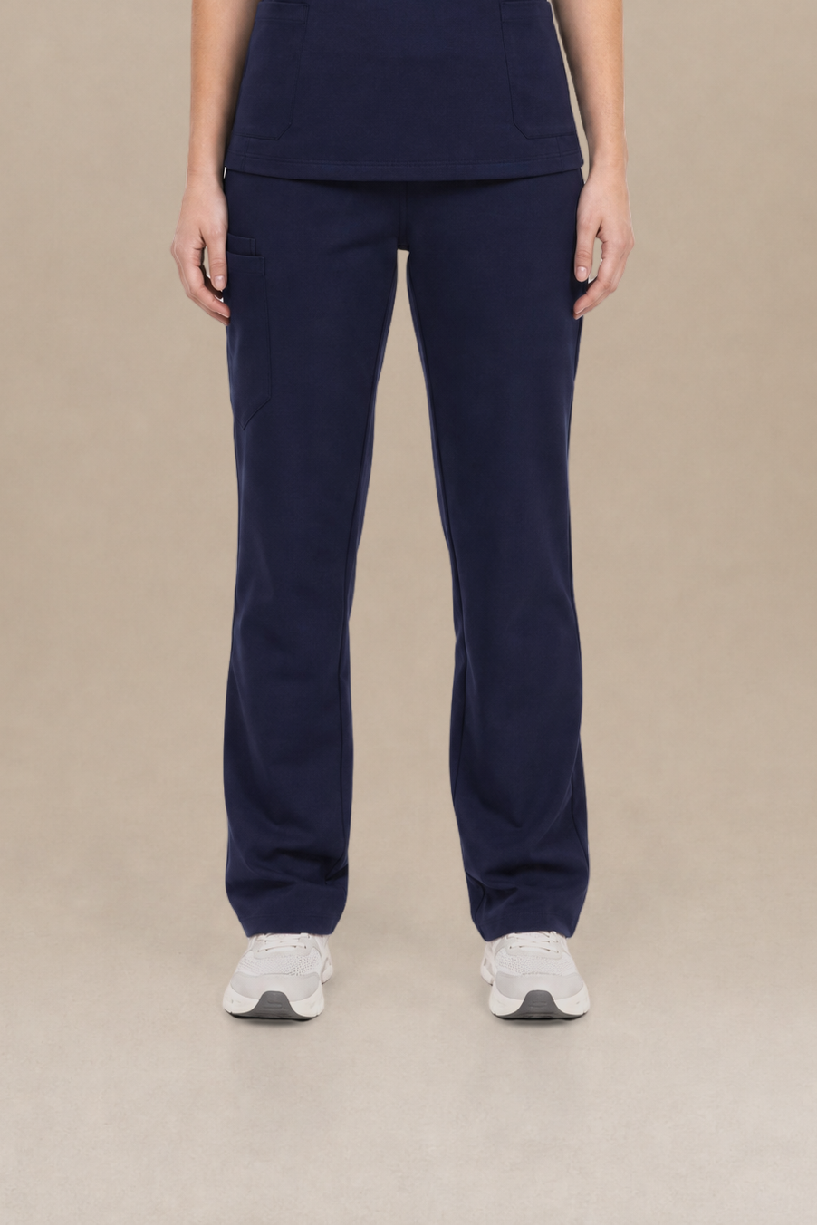 A woman modeling a matching navy blue scrub set consisting of a short-sleeve V-neck top and straight-leg pants, standing against a solid beige studio background.