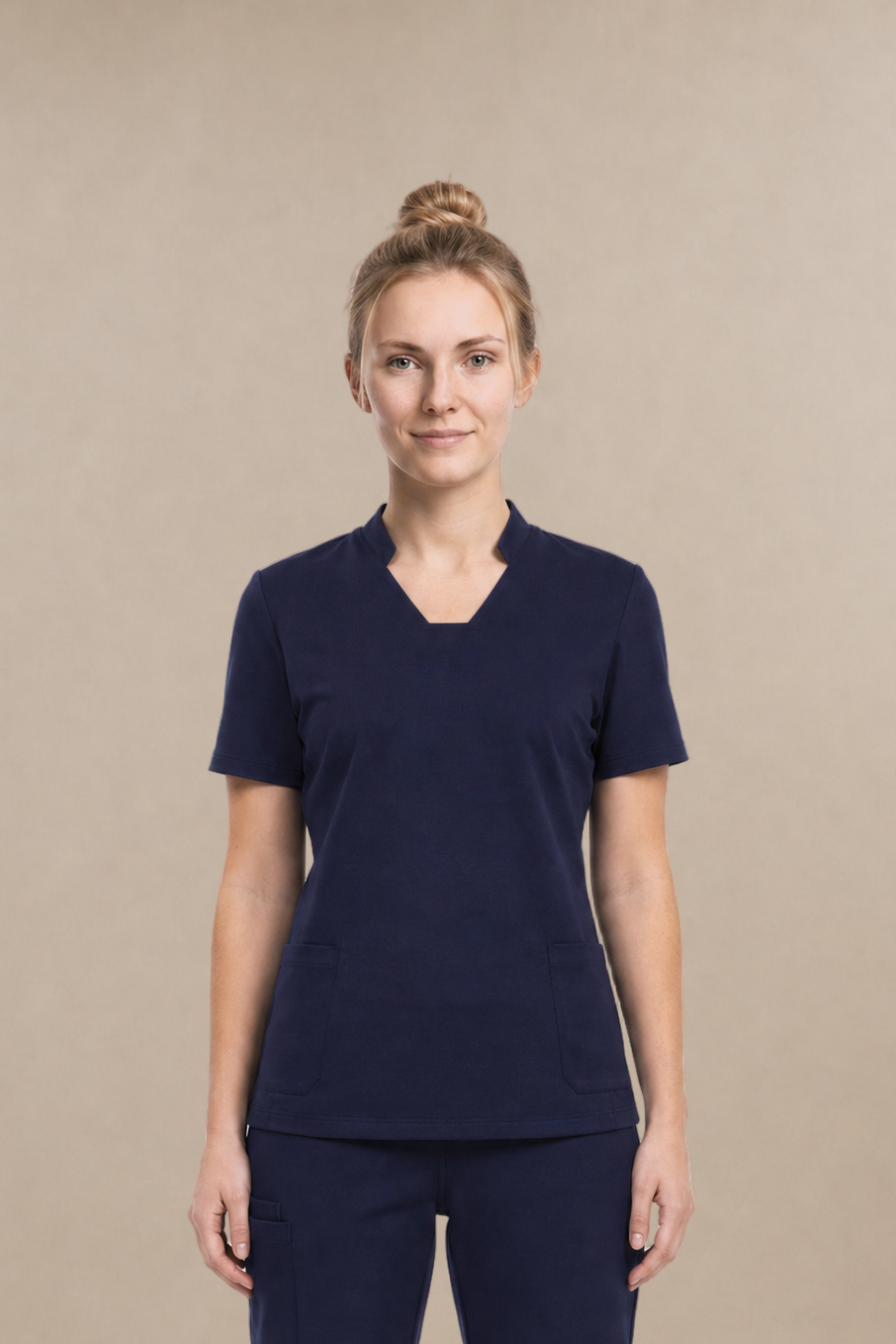 A woman modeling a matching navy blue scrub set consisting of a short-sleeve V-neck top and straight-leg pants, standing against a solid beige studio background.
