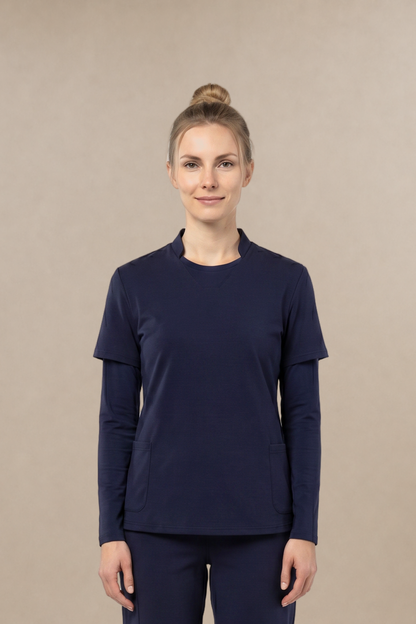 A woman modeling a matching navy blue long-sleeve scrub top and pants set with an underscrub top, standing against a solid beige studio background.