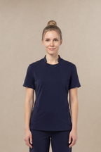 A woman stands facing forward wearing a matching navy blue short-sleeve crewneck underscrub top layered under a navy blue short-sleeve scrub top and pants scrub set, against a plain beige studio background.