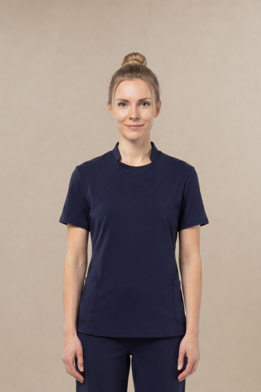 A woman stands facing forward wearing a matching navy blue short-sleeve crewneck top and pants scrub set, paired with white sneakers, against a plain beige studio background.