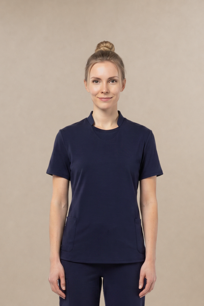 A woman stands facing forward wearing a matching navy blue short-sleeve crewneck top and pants scrub set, paired with white sneakers, against a plain beige studio background.