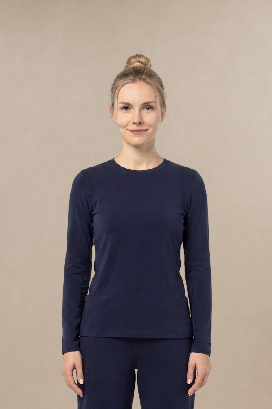 A woman stands facing forward in a matching navy long-sleeve underscrub top and navy scrub pants, with white sneakers, against a plain warm beige studio background.