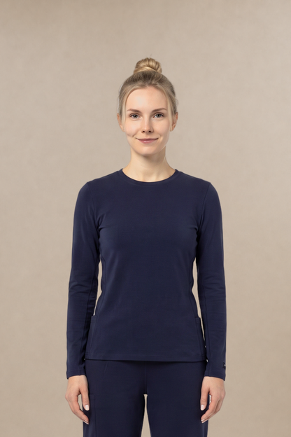 A woman stands facing forward in a matching navy long-sleeve underscrub top and navy scrub pants, with white sneakers, against a plain warm beige studio background.