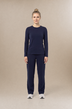 A woman stands facing forward in a matching navy long-sleeve underscrub top and navy scrub pants, with white sneakers, against a plain warm beige studio background.