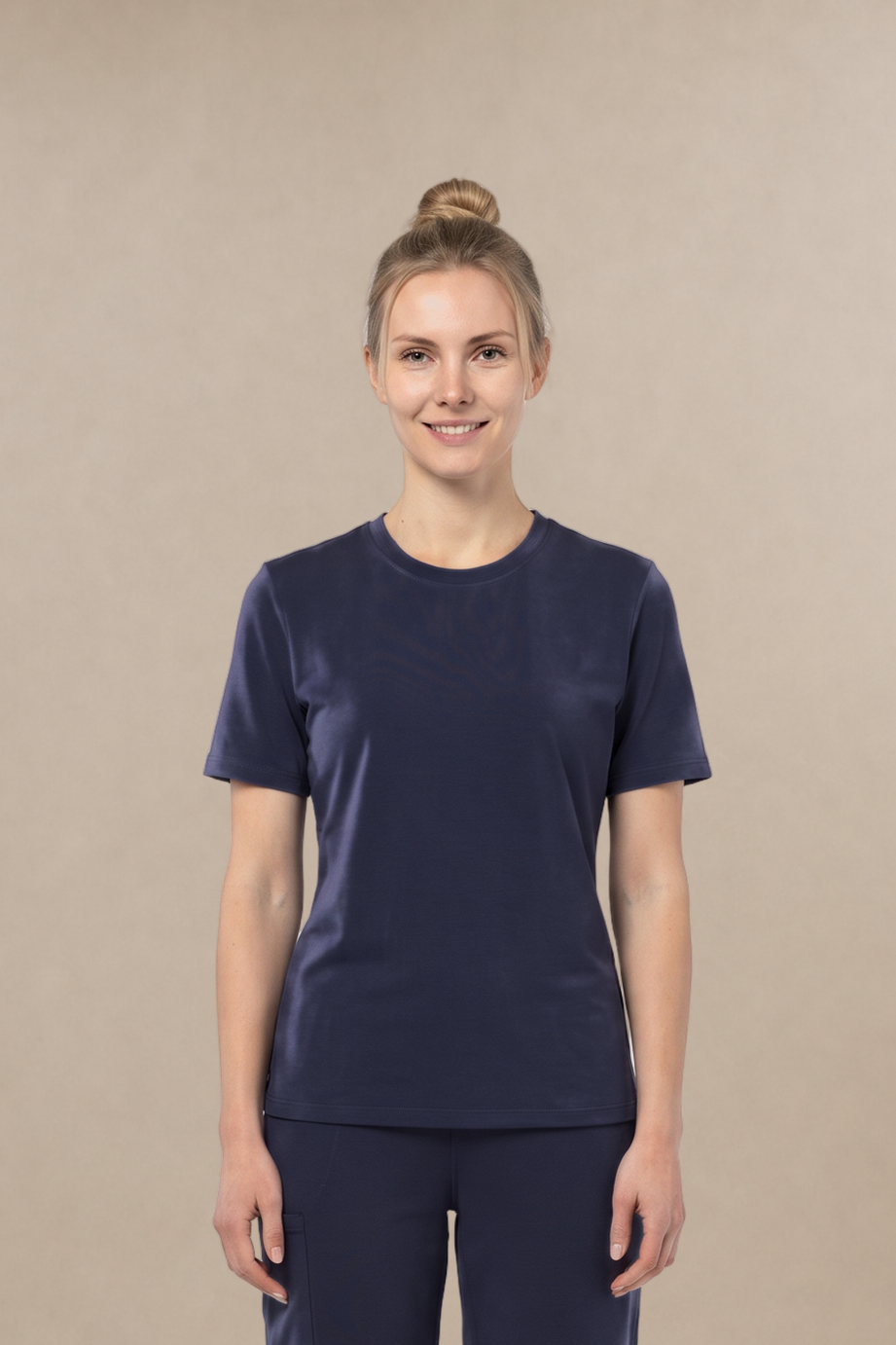 A woman modeling a matching navy blue scrub set, consisting of a short-sleeve crewneck underscrub top and straight-leg pants, standing against a solid beige studio background.