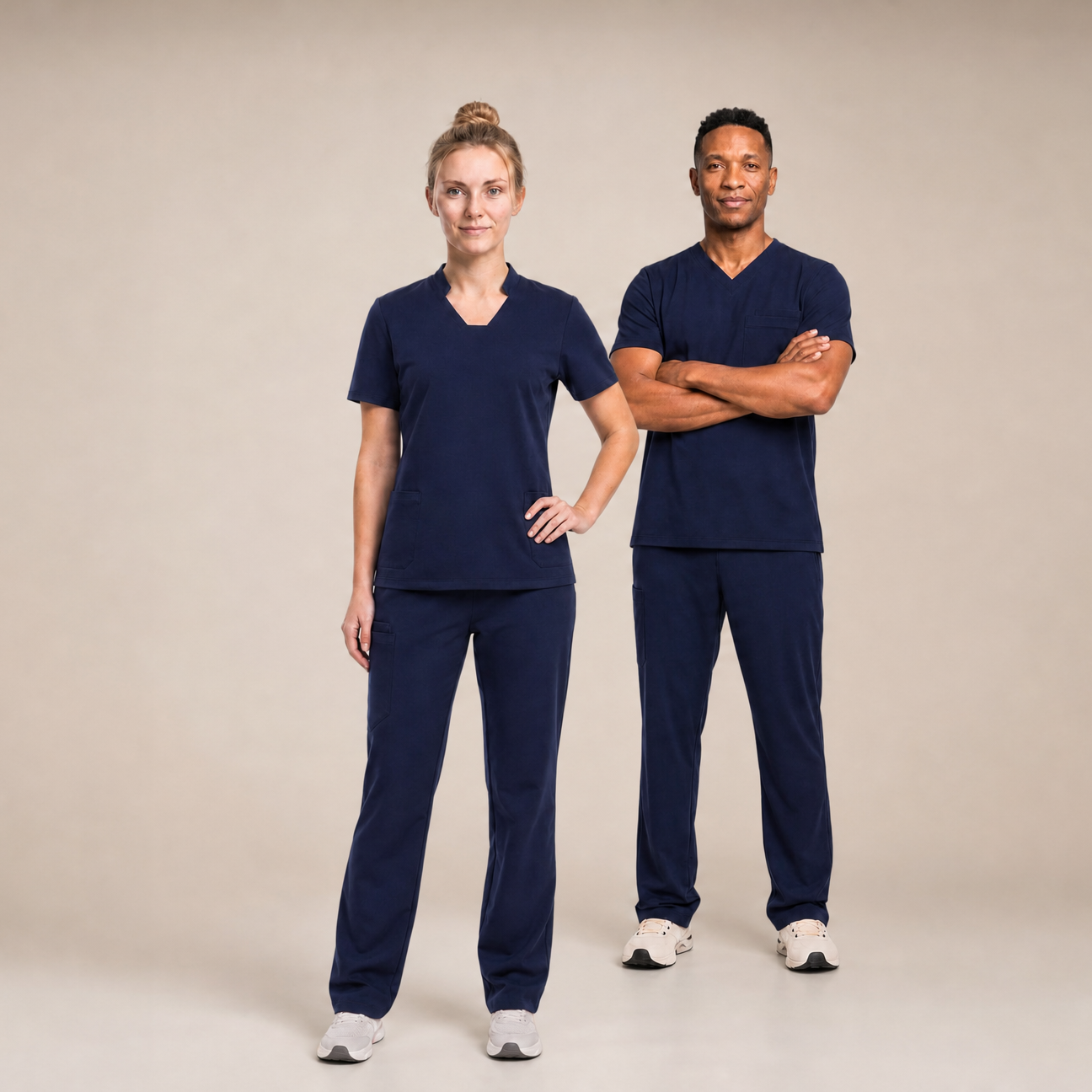 A man and a woman modeling matching navy blue V-neck scrub sets against a plain beige studio background.