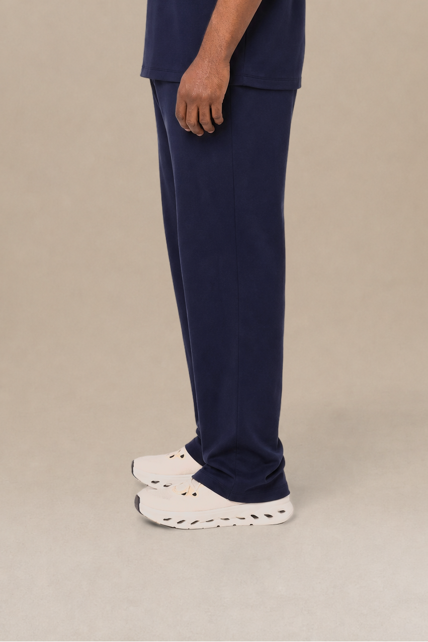 A man in profile wearing a navy blue short-sleeve t-shirt and matching navy blue scrub pants, standing against a plain beige studio background.