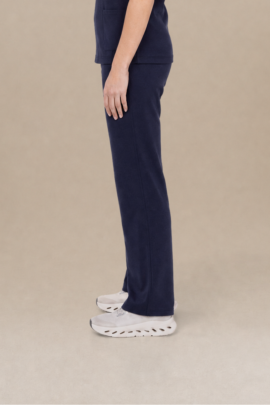 A woman in a side profile wearing a navy blue scrub set consisting of a short-sleeve top and matching pants, paired with white sneakers, against a solid beige studio background.