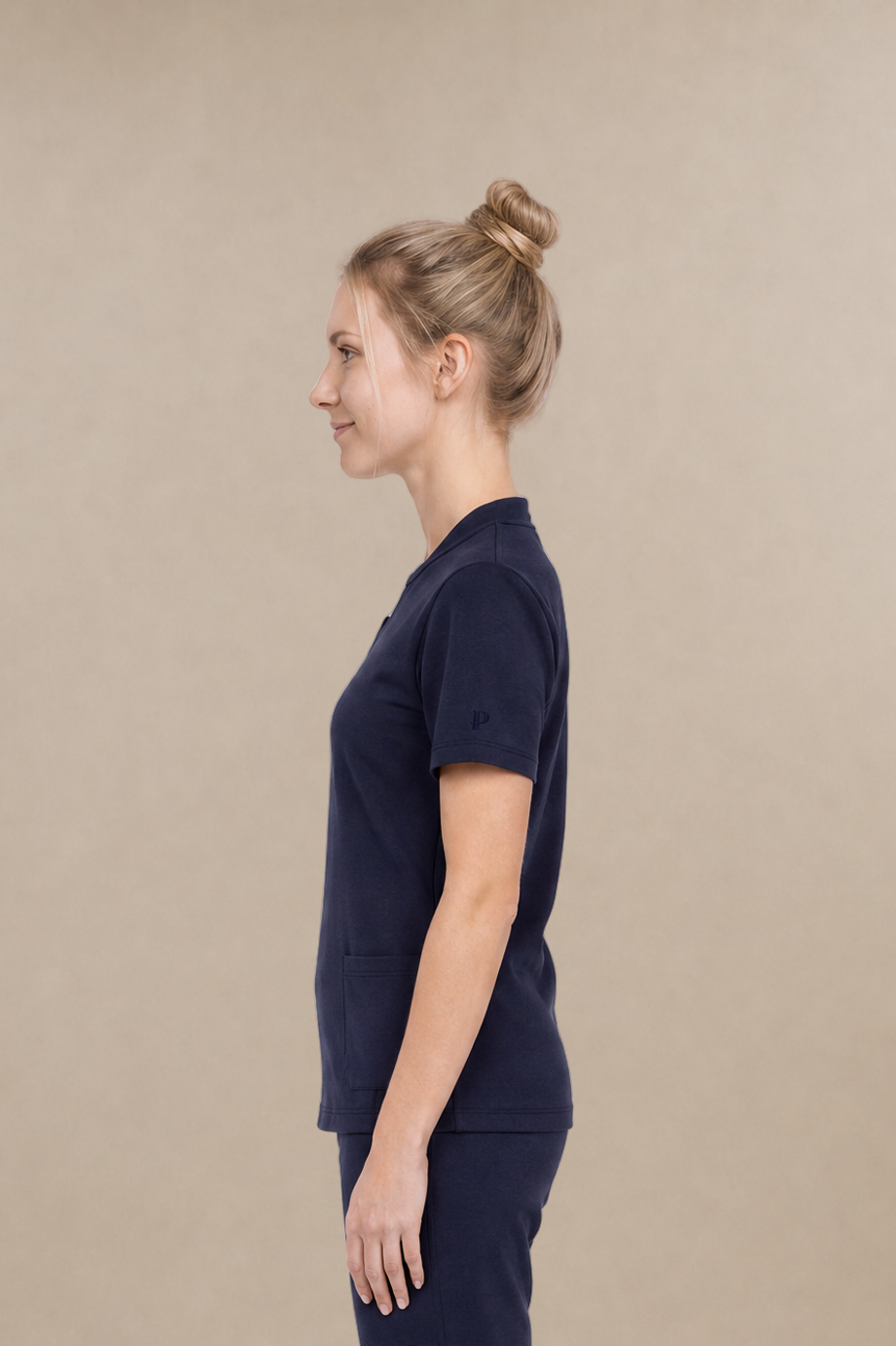 A woman in a side profile wearing a navy blue scrub set consisting of a short-sleeve top and matching pants, against a solid beige studio background.