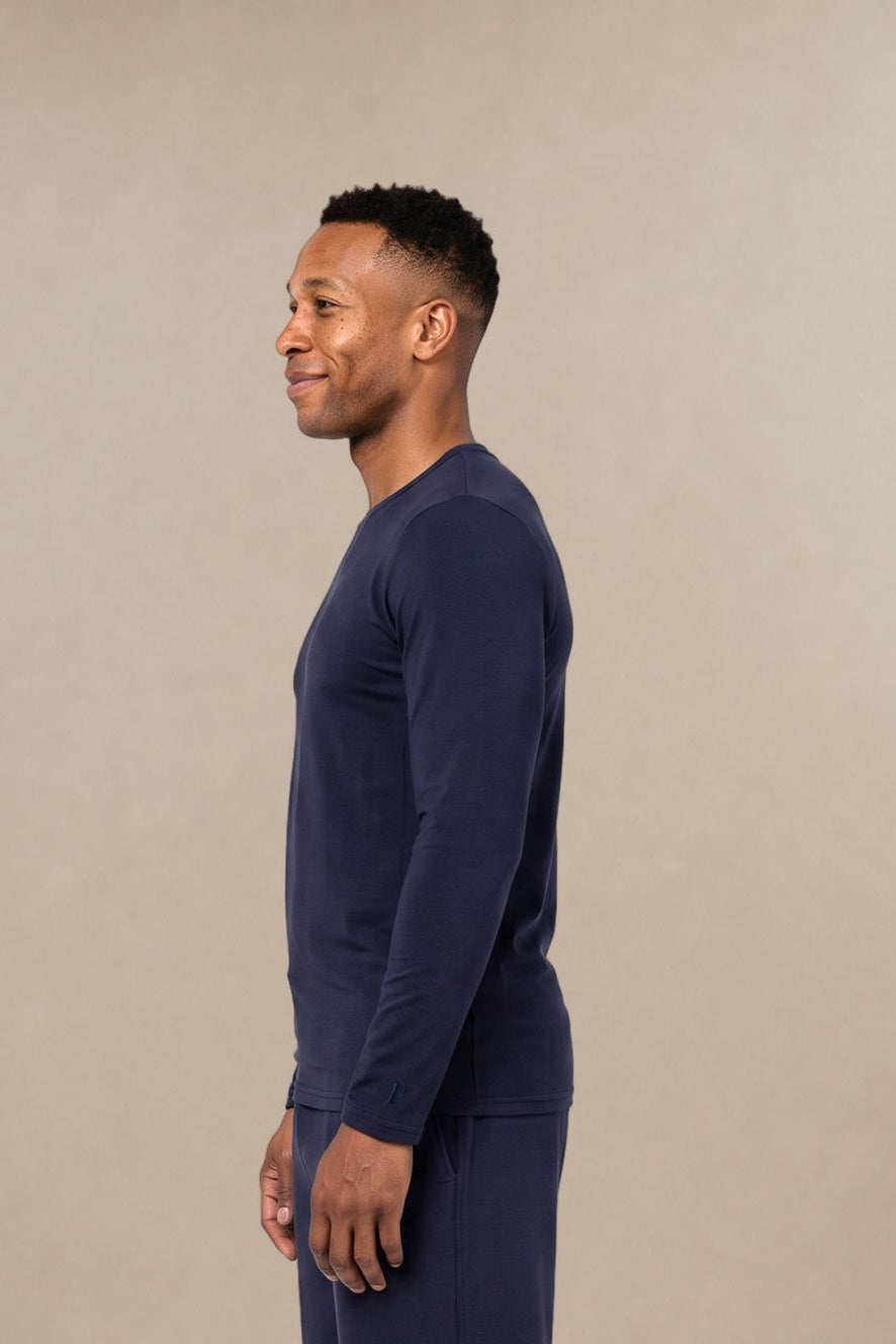 A man in profile wearing a navy blue long-sleeve crewneck subscrub shirt and matching navy blue pants, standing against a plain beige studio background.