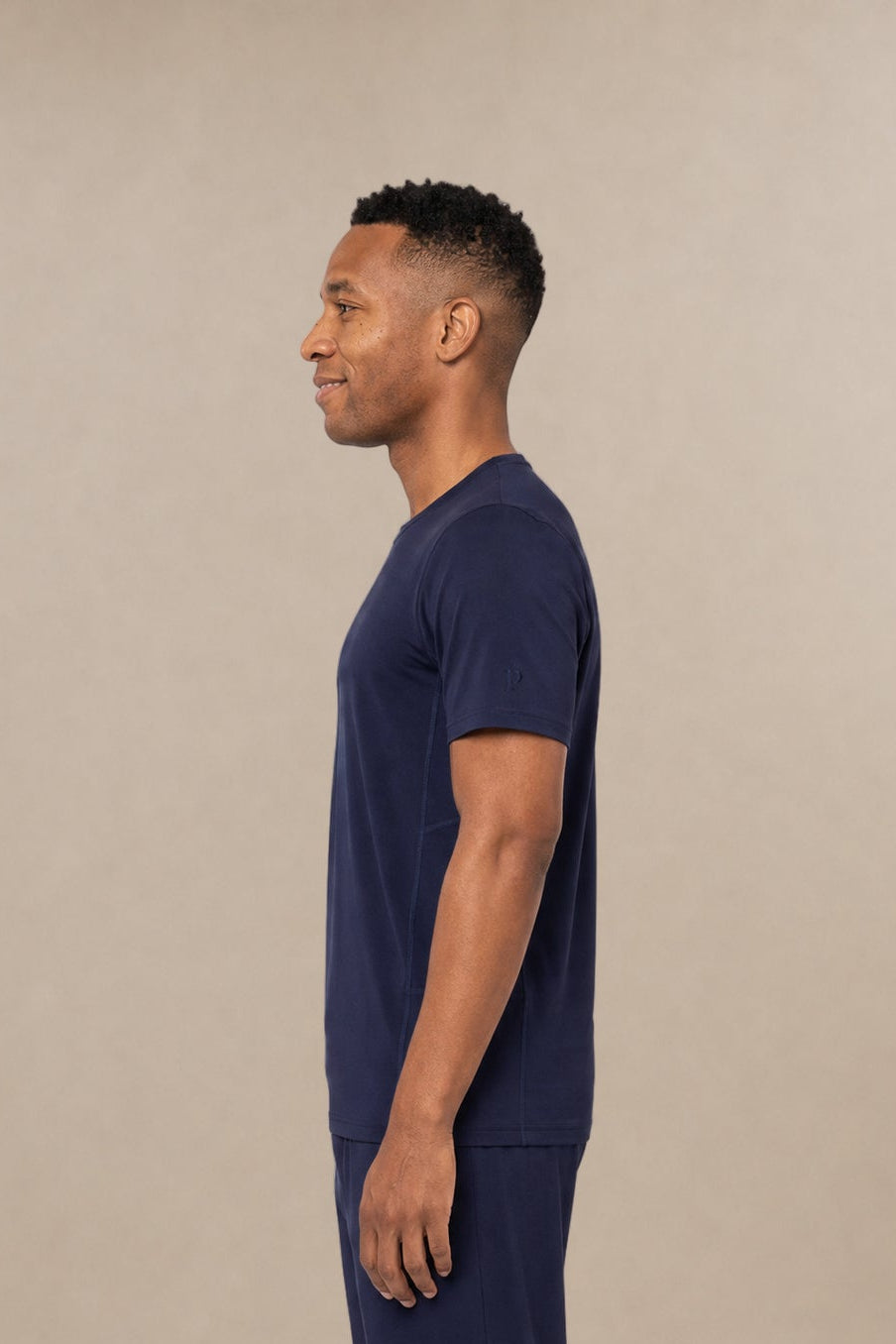 A man in profile wearing a navy blue short-sleeve subscrub shirt and matching navy blue scrub pants, standing against a solid beige studio background.