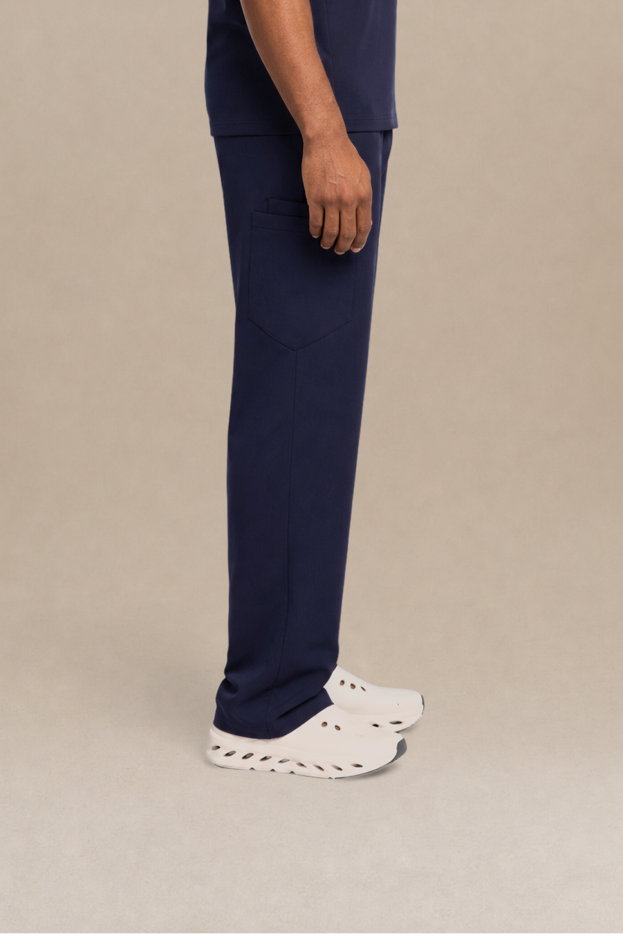 A man in profile wearing a navy blue short-sleeve V-neck t-shirt and matching navy blue scrub pants, paired with white sneakers, standing against a solid beige studio background.