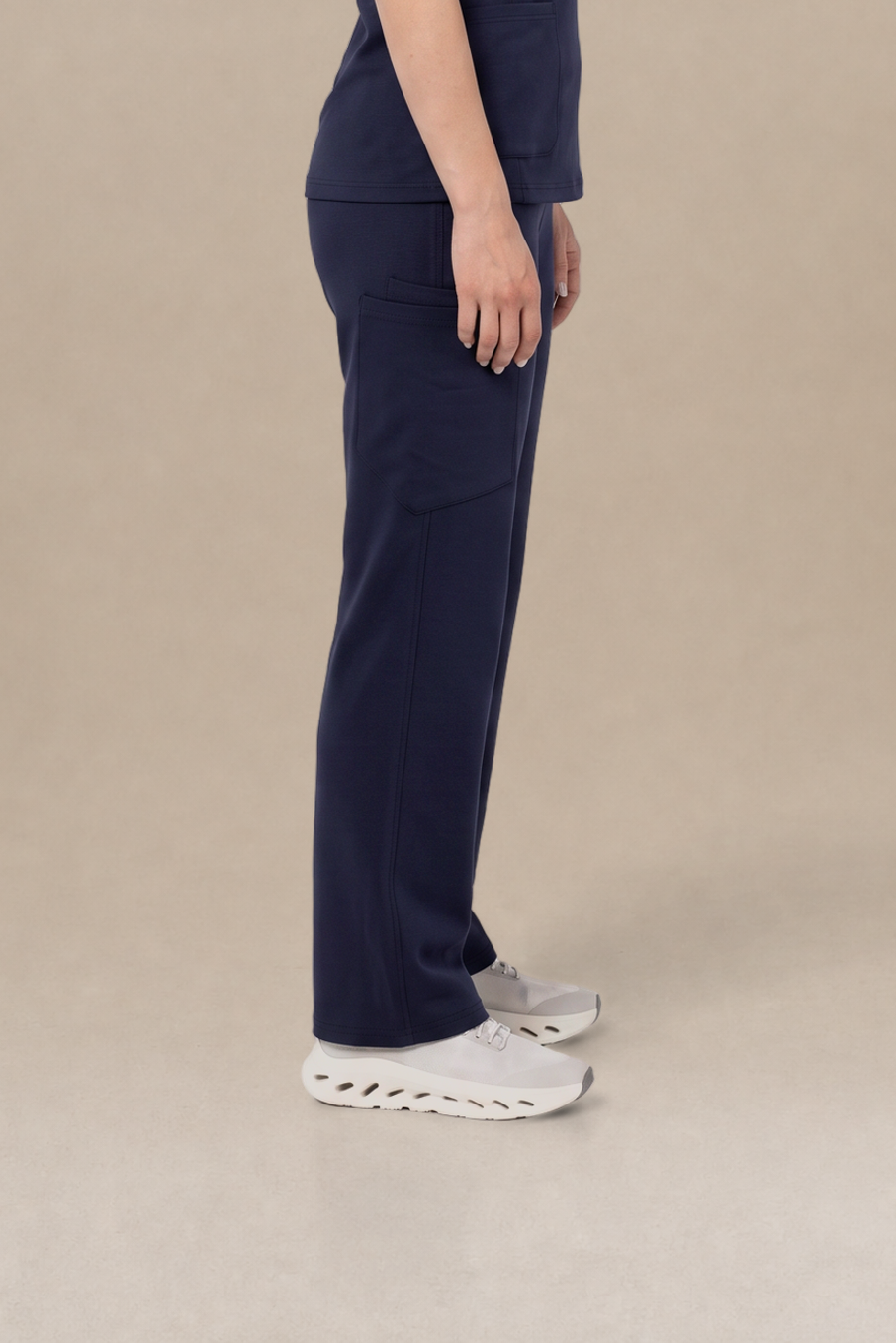 A woman stands in profile wearing a dark navy blue scrub set consisting of a short-sleeve top and matching pants, paired with white sneakers, against a plain beige studio background.