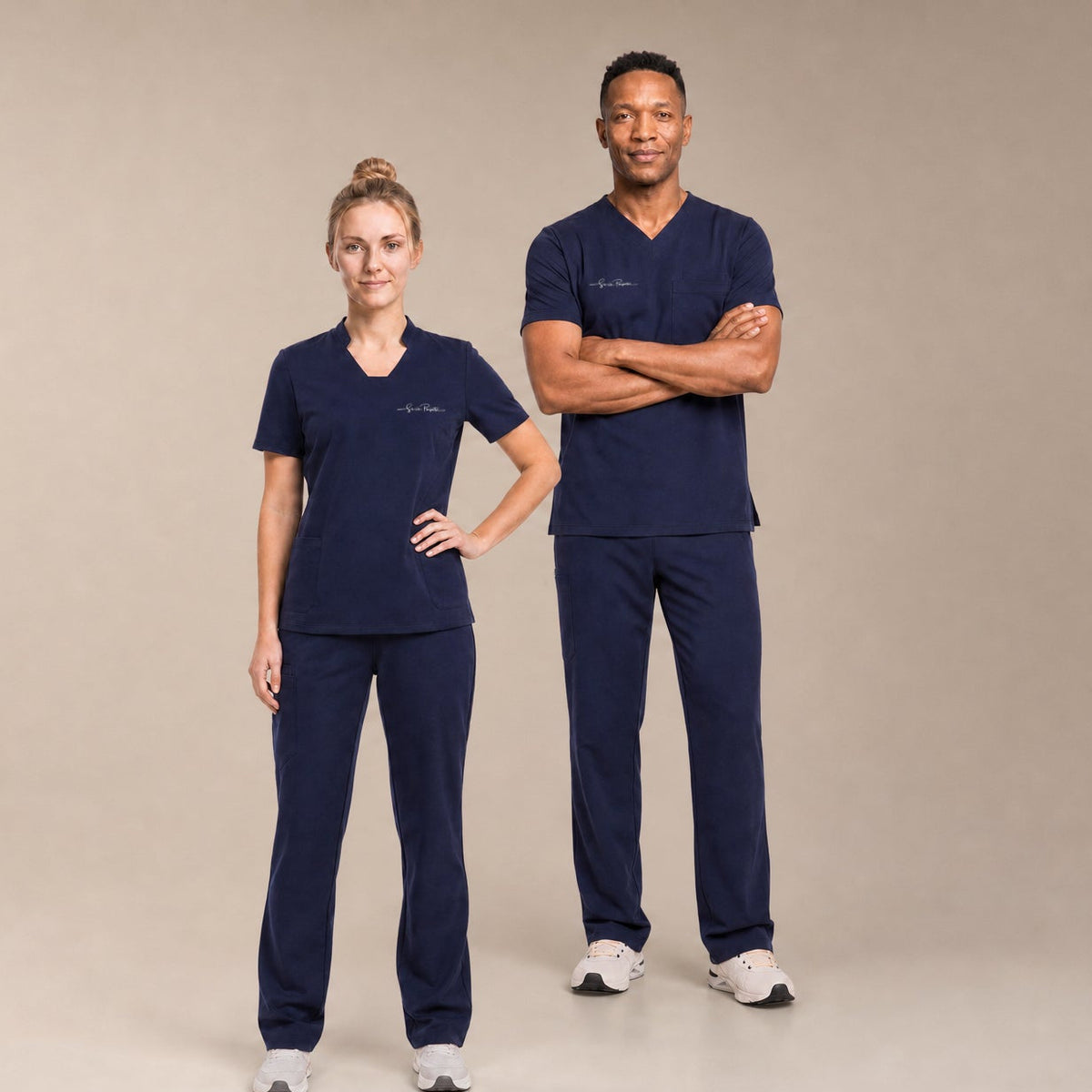Two healthcare workers, a woman and a man, standing in matching dark navy blue scrub sets against a solid beige studio background.
