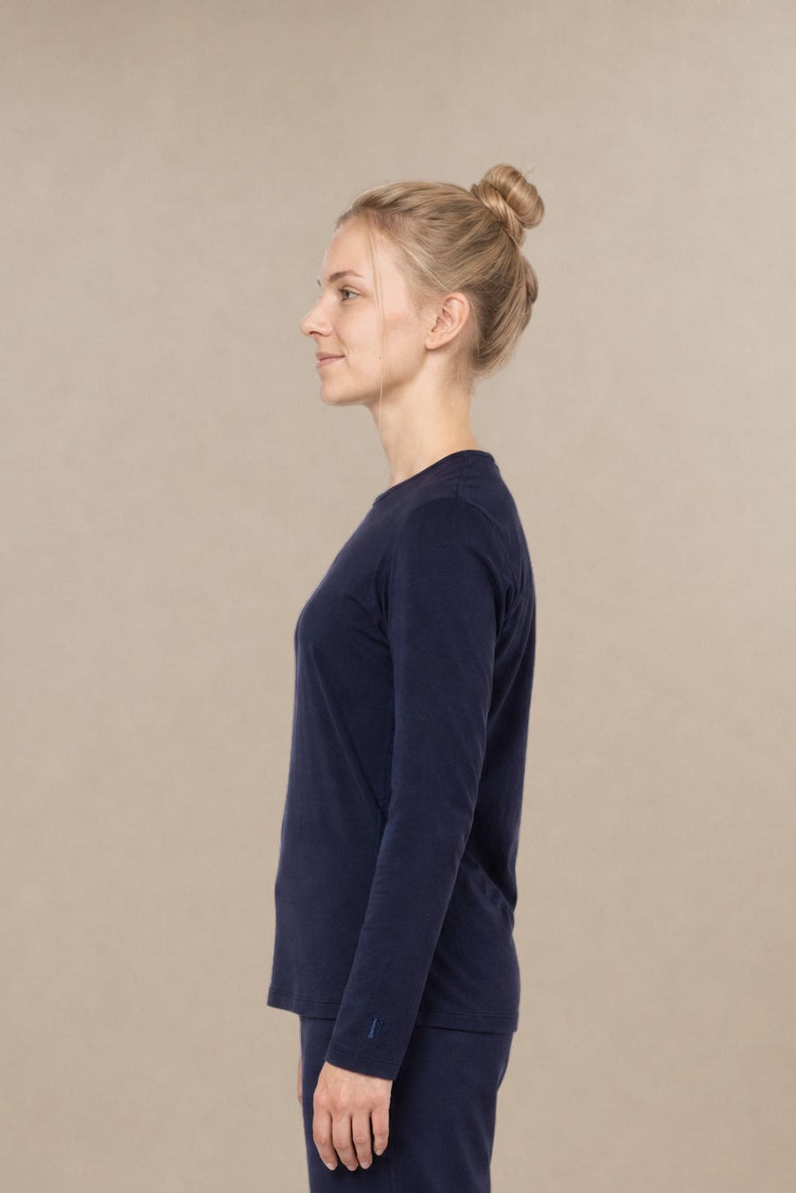 Woman in profile view wearing a navy blue, long-sleeve scrub top and matching straight-leg scrub bottoms against a beige background.