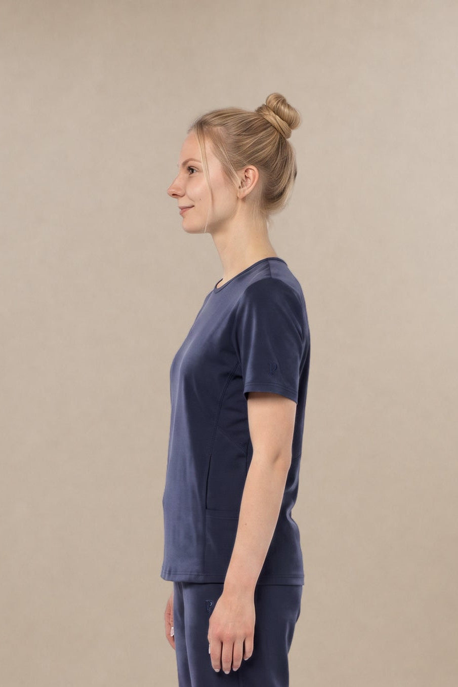 A woman in a side profile wearing a matching navy blue short-sleeve scrub top and wide-leg scrub pants set, paired with white sneakers, against a plain beige studio background.